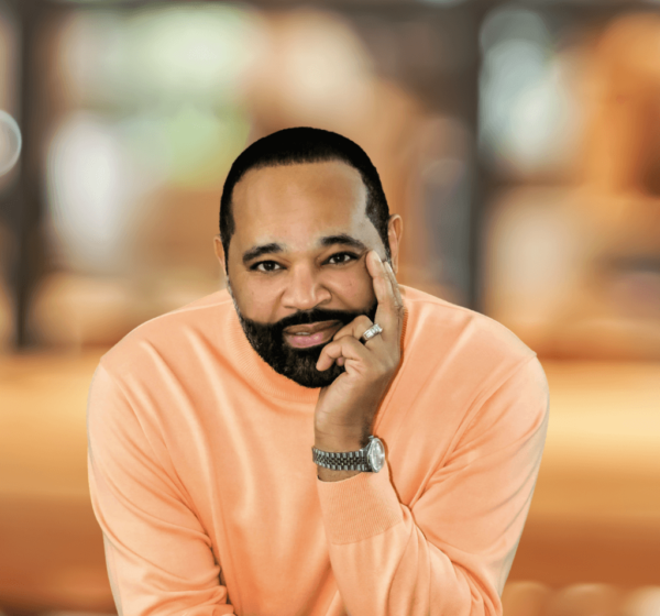 A man with beard and mustache sitting in an orange sweater.